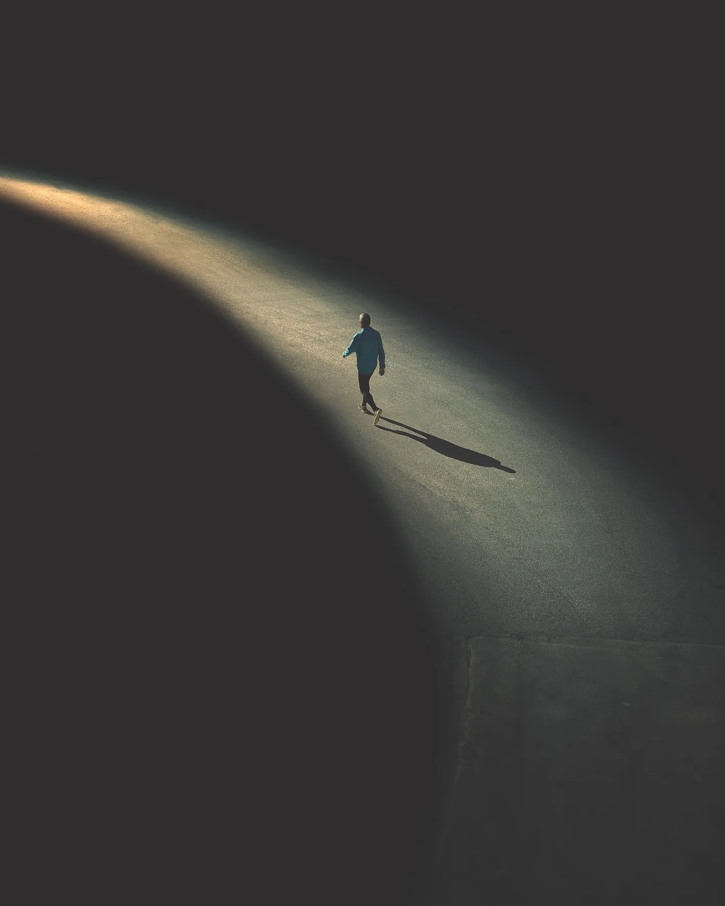 A lone figure in a blue shirt walks along a narrow band of sunlight that cuts across dark pavement.